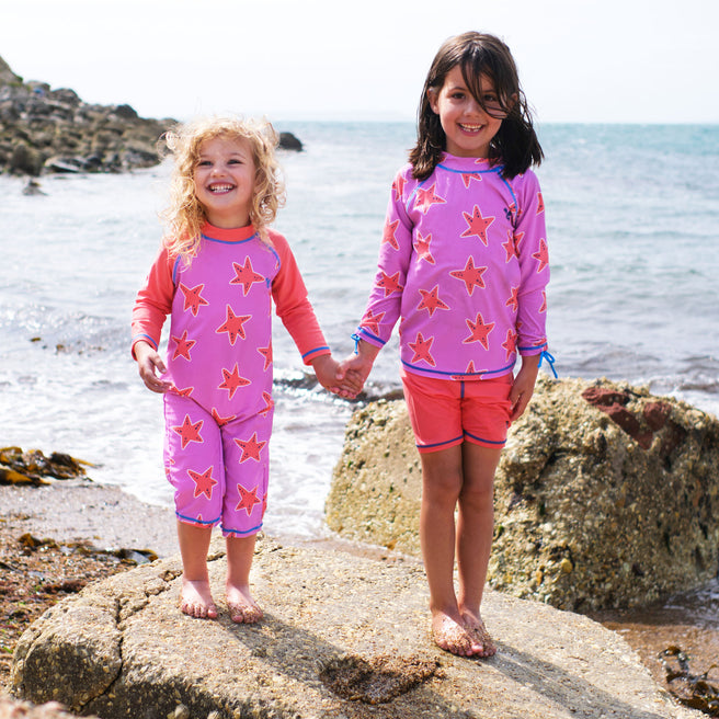 Two children wearing UV Protective Surf Set Pink Starfish, standing on a rock on a beach, smiling at the camera.